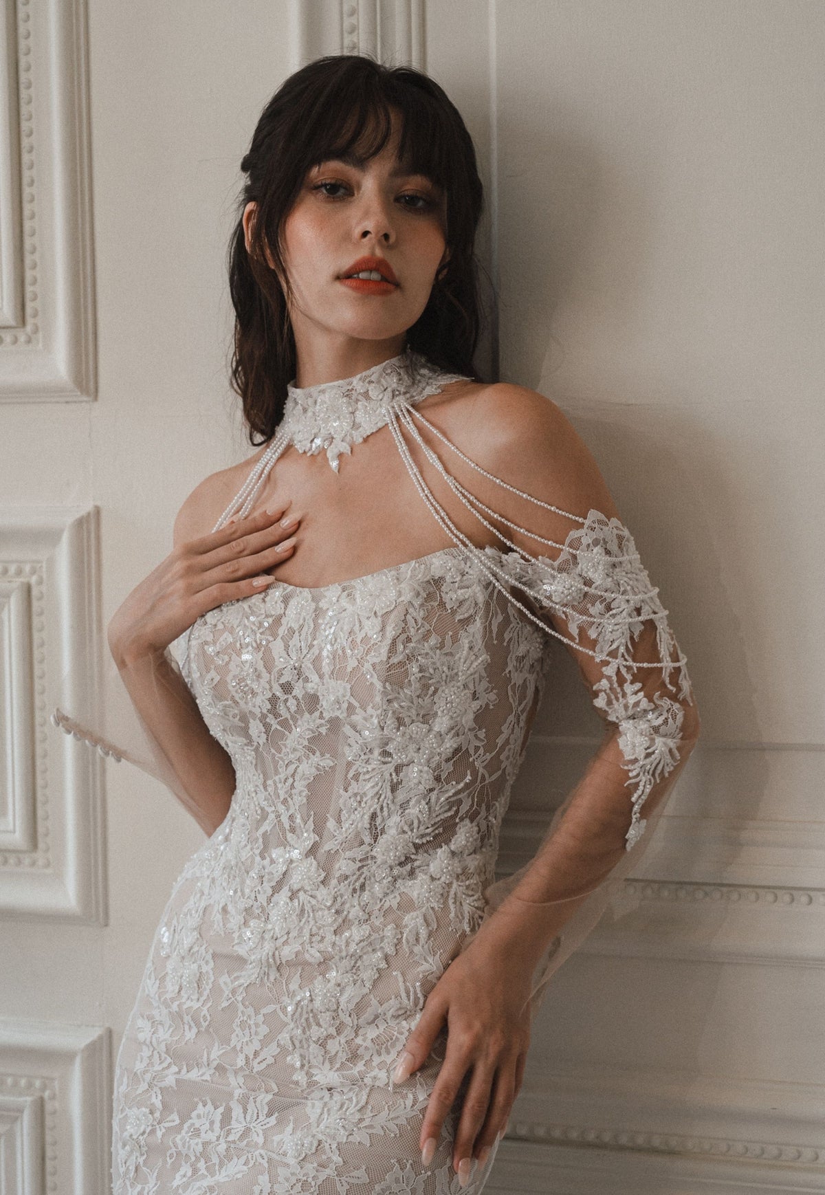 A woman wearing a white lace bridal gown with a high lace collar, strappy shoulder details, and a fitted silhouette, posed against an off-white paneled wall.
