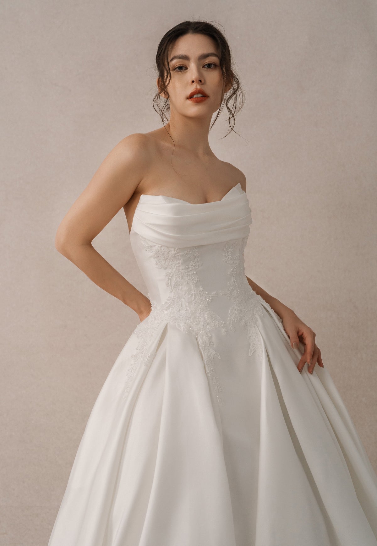 A woman modeling a strapless white wedding gown with a gathered sweetheart neckline, lace-embellished bodice, and full satin skirt against a soft beige studio background.