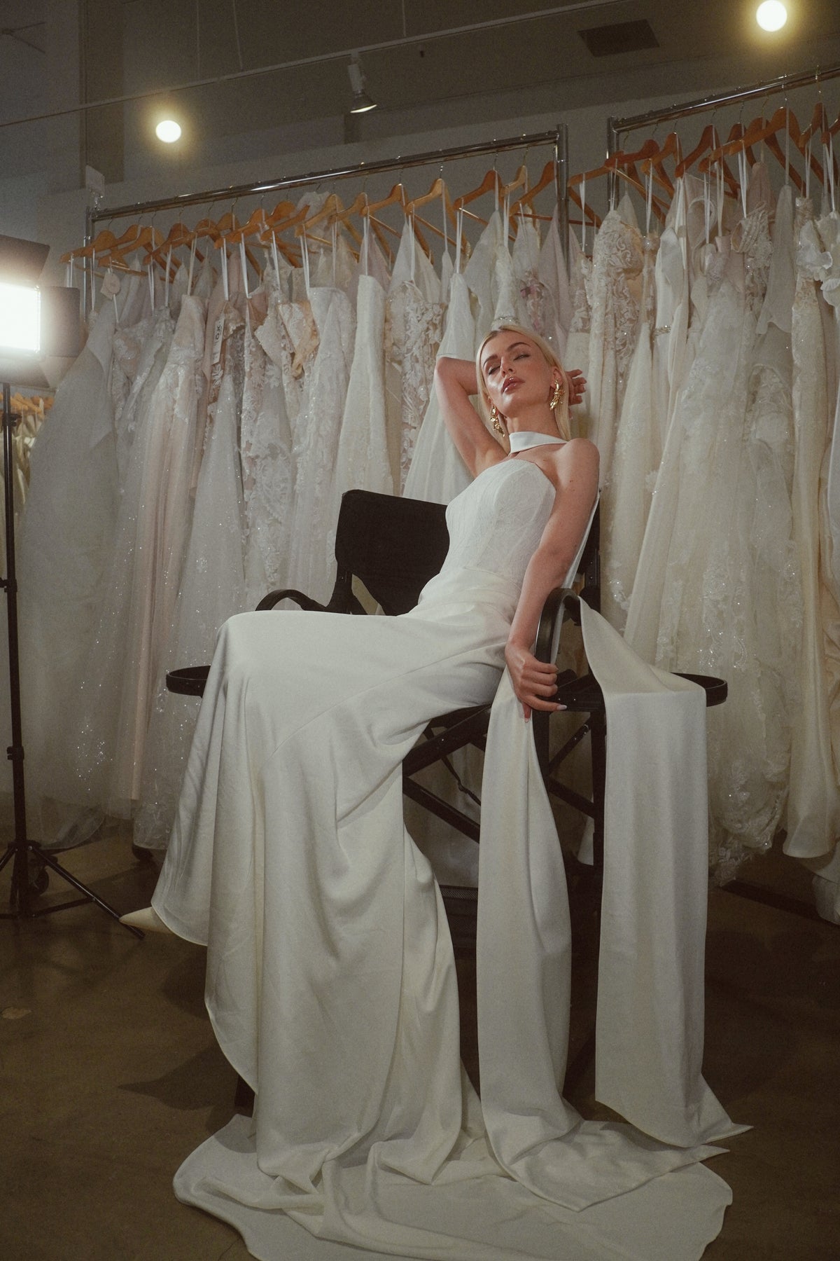 A woman wearing a sleeveless white bridal gown sits on a black chair in a bridal boutique, with rows of hanging wedding dresses in the background.