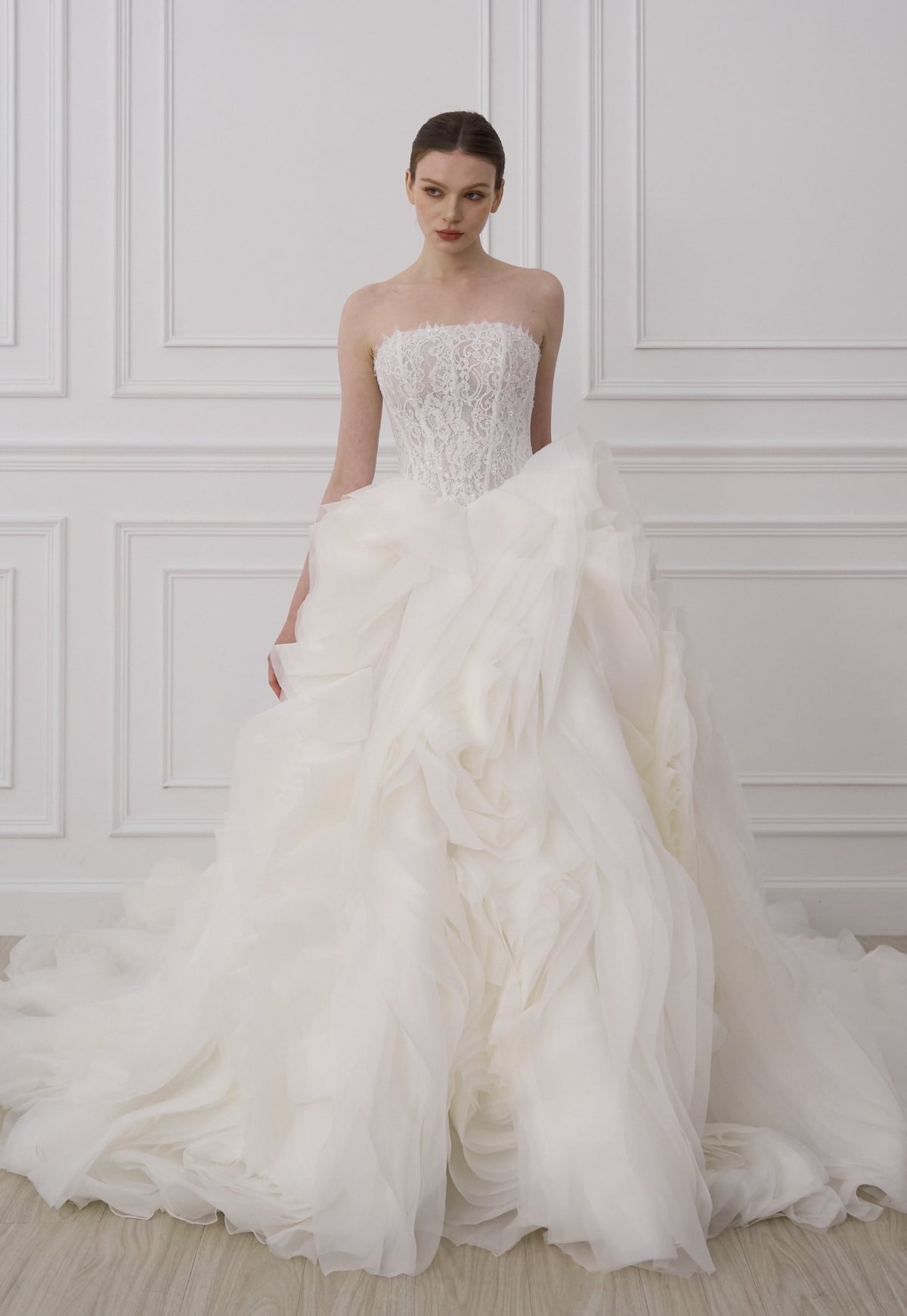 A woman modeling a strapless white wedding gown with a fitted lace bodice and a voluminous layered tulle skirt, standing in a bright room with white paneled walls and light wood flooring.